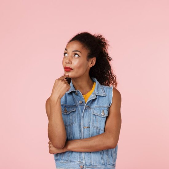 Woman standing in front of a pink wall in a thinking pose, pondering the most common vegan questions and how to respond to them.