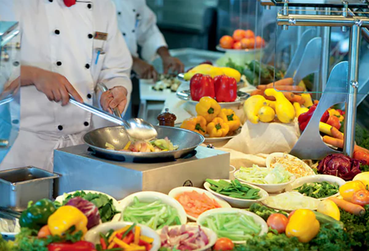 A chef in a white uniform stir frying vegetables next to a table laden with fresh sliced fresh veggies and fruit. 
