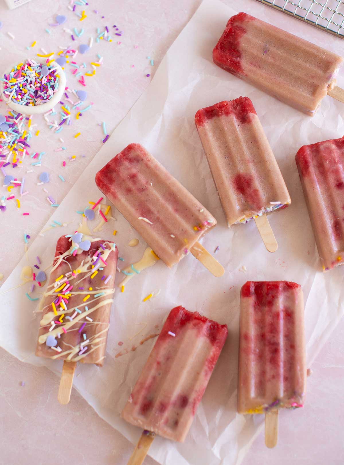 Creamy strawberry popsicles laid out on parchment paper with a drizzle of vegan white chocolate. 