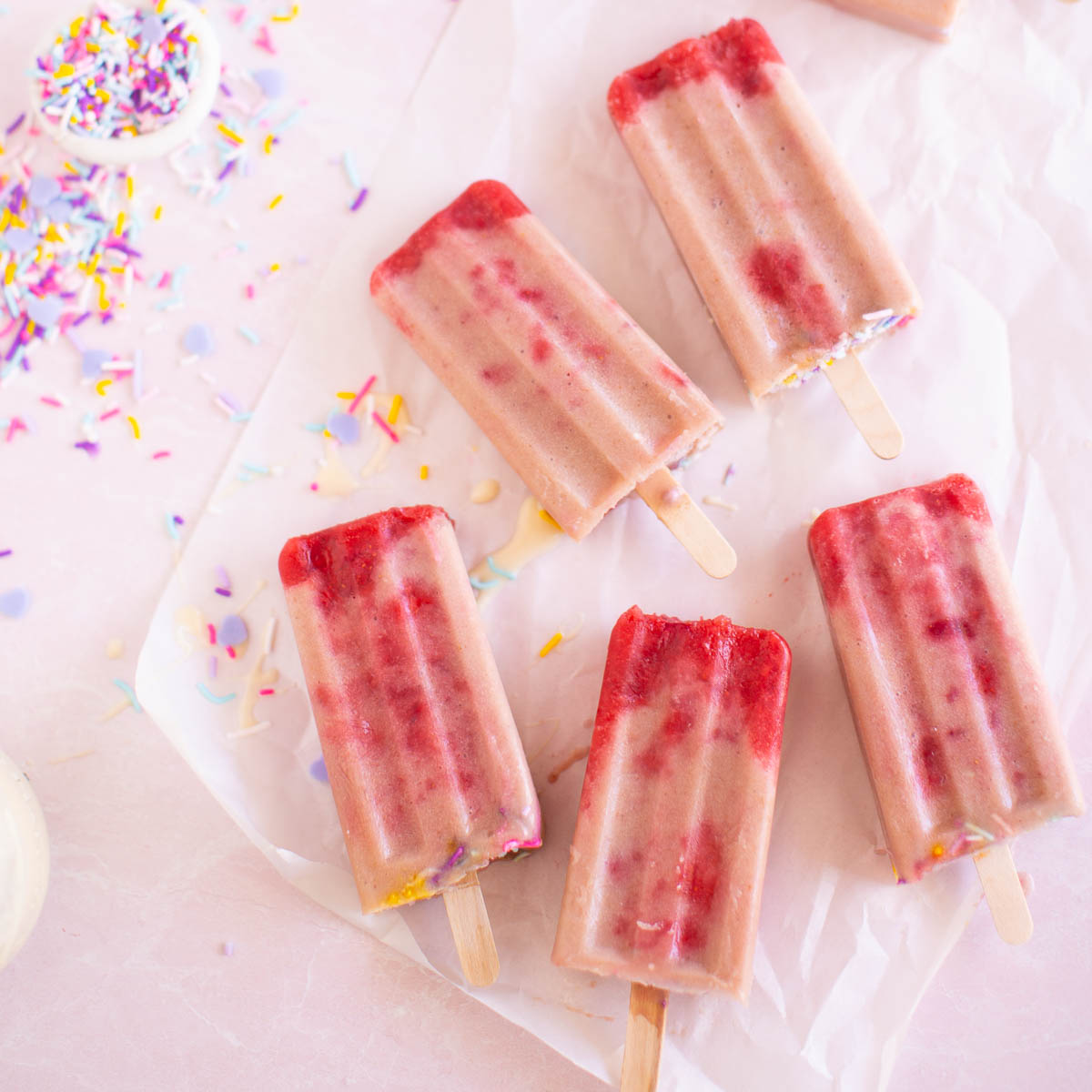 A flatlay of homemade strawberry banana popsicles on a pink table with colorful sprinkles.