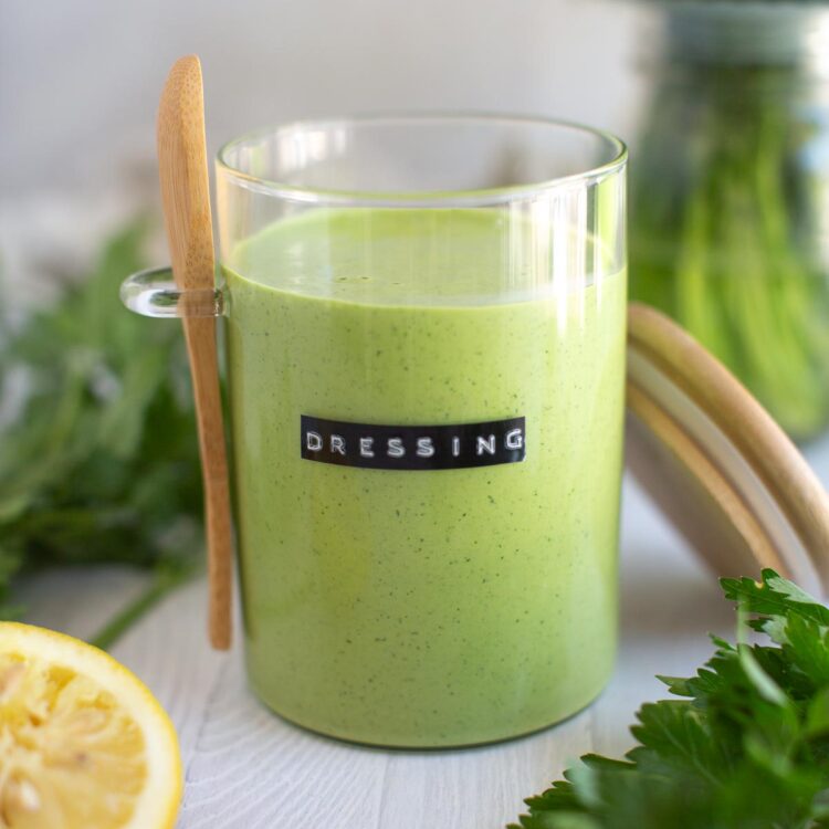 A glass jar filled with vegan green goddess dressing, surrounded by fresh herbs and lemon slices.