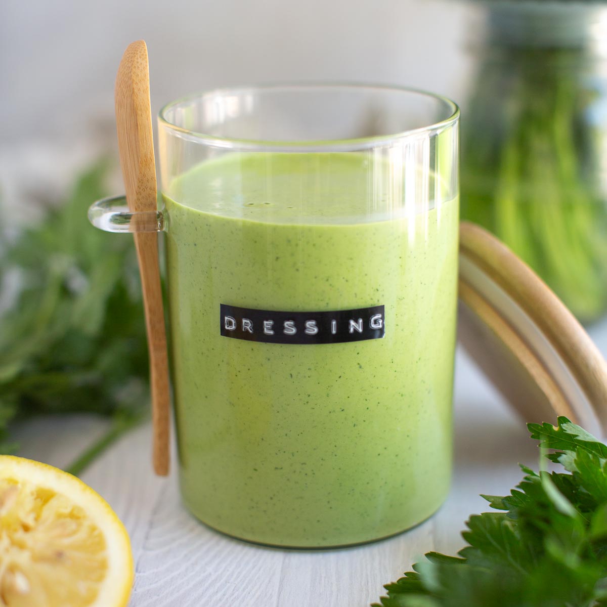 A glass jar filled with vegan green goddess dressing, surrounded by fresh herbs and lemon slices.