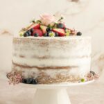 Finished vegan sponge cake topped with berries and a rose on a white cake stand.
