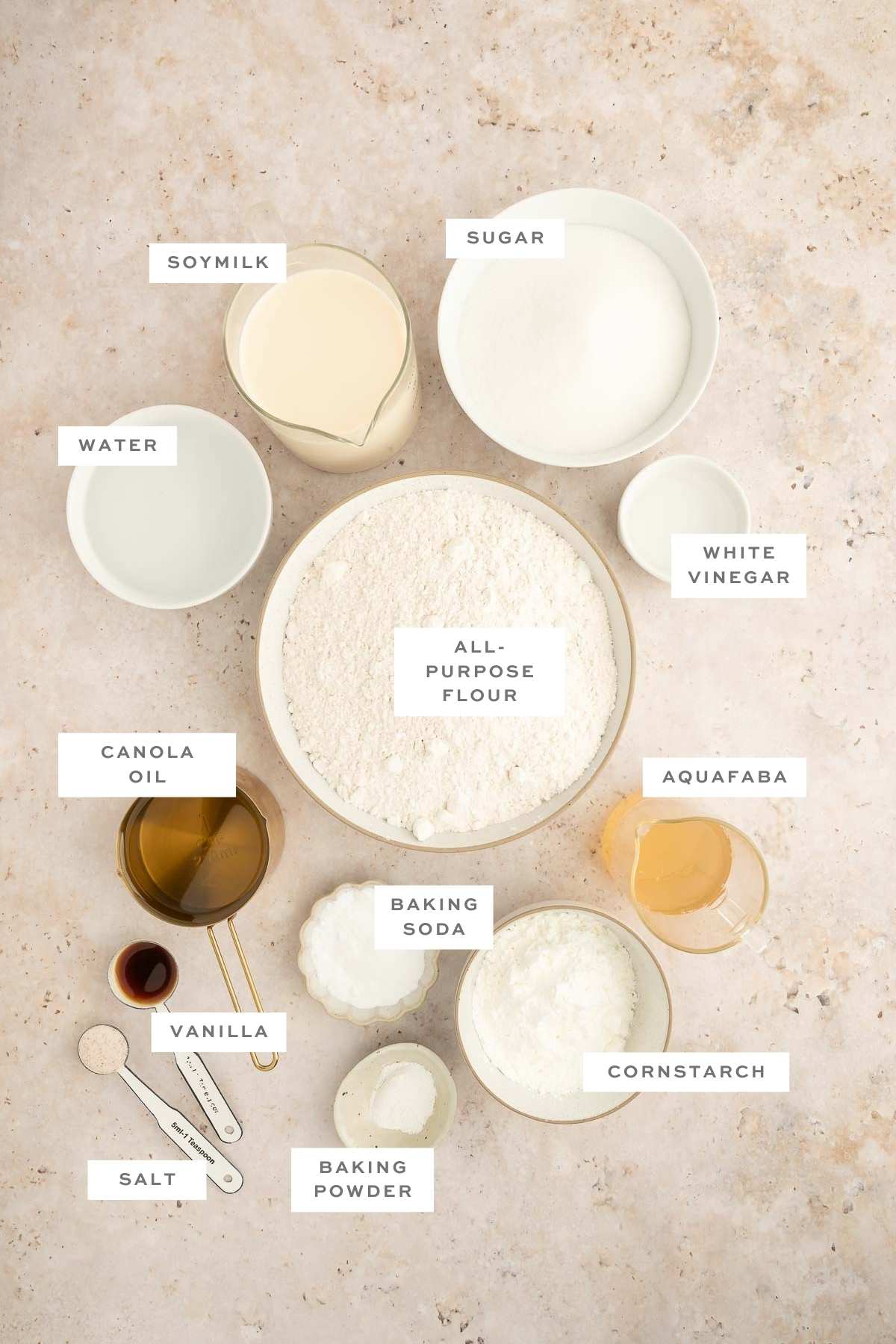 Flat lay of labeled ingredients for vegan sponge cake, including flour, aquafaba, canola oil, and soy milk.