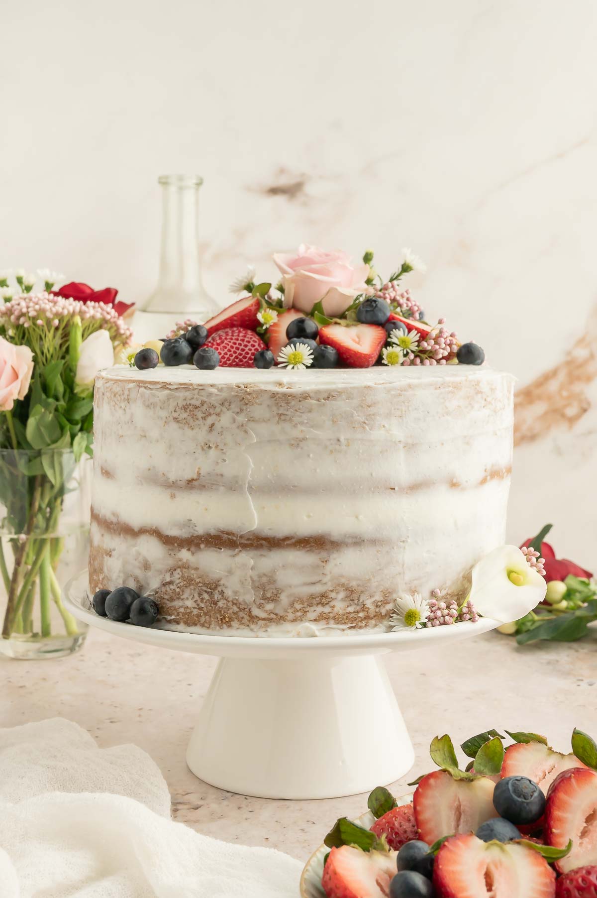 Fully frosted vegan sponge cake topped with berries and florals, styled with fresh flowers in the background.