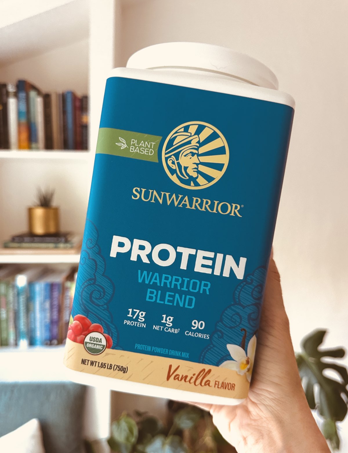 A container of SunWarrior vegan vanilla protein powder, warrior blend, held up in a well lit room.