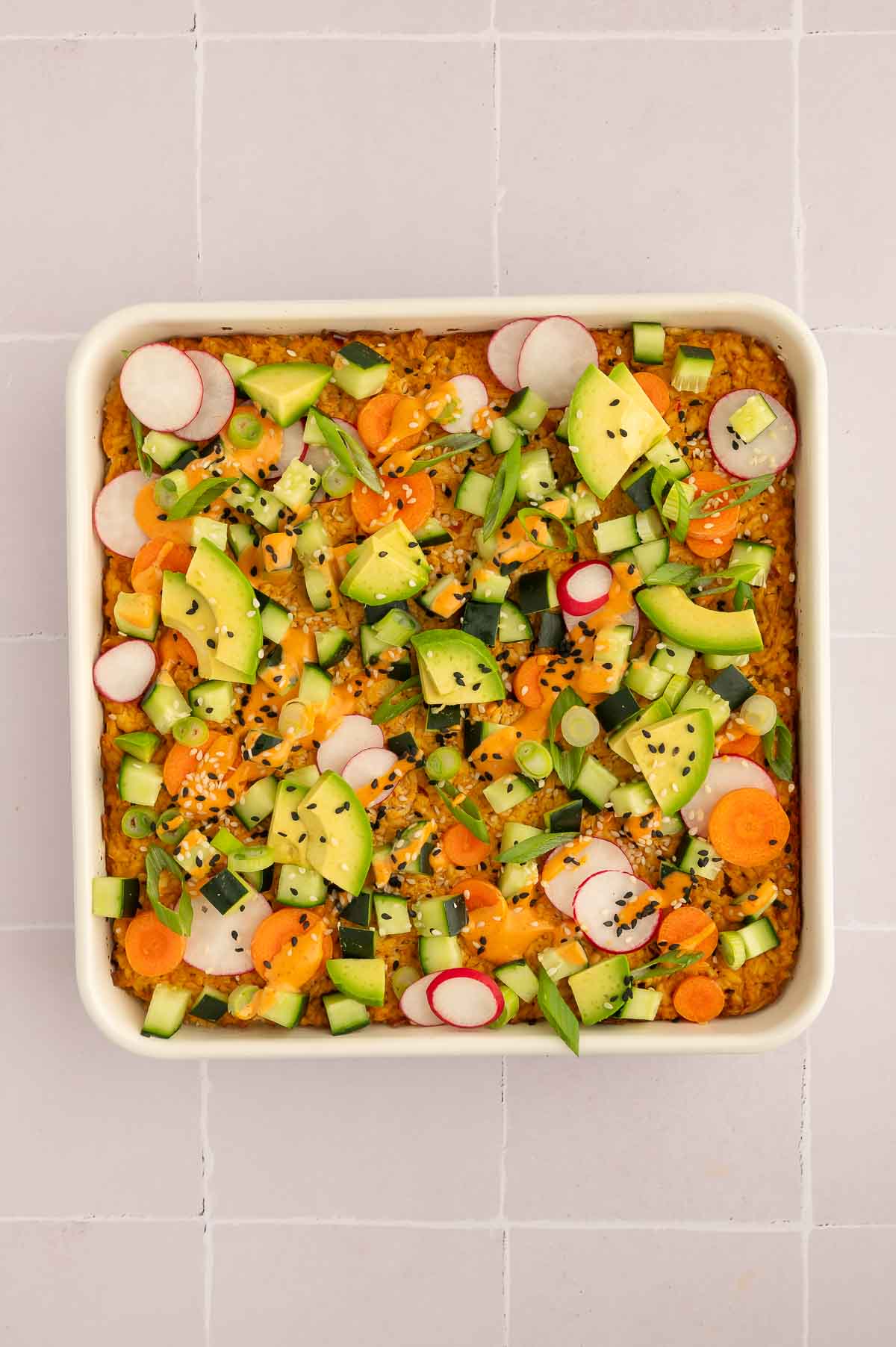 Vegan sushi bake in a baking dish topped with sliced radish, avocado, spicy mayo, cucumber, carrots, and sesame seeds.