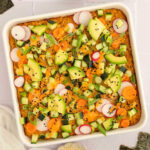 A casserole dish filled with vegan sushi bake and topped with fresh sliced veggies, avocado cubes, and sesame seeds.
