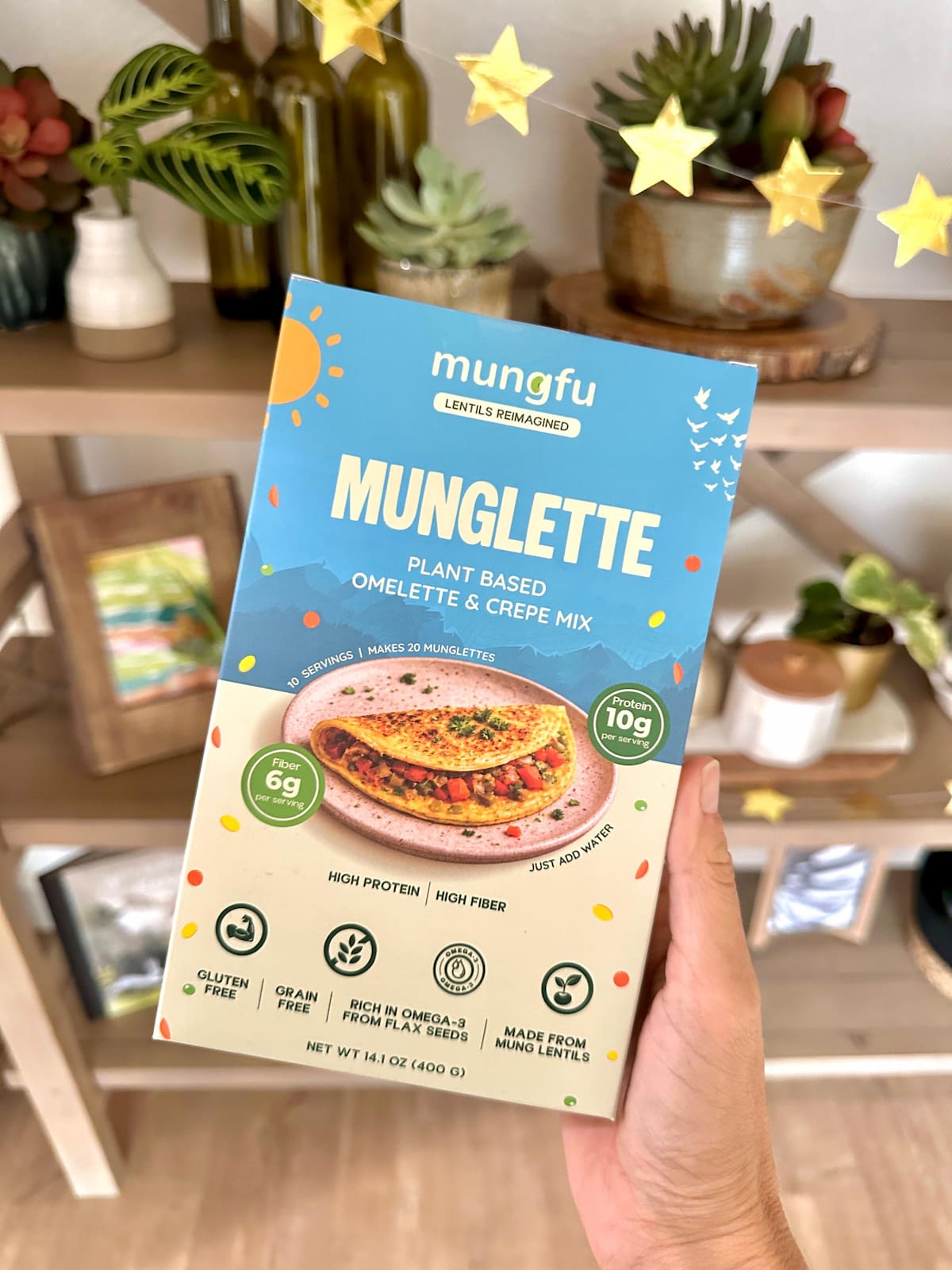 A hand holding a blue and cream package of munglette lentil-based omlette and crepes mix.