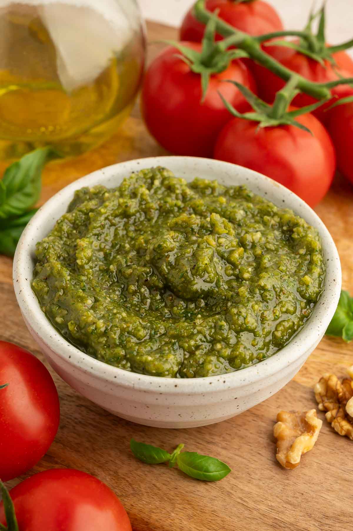 Basil walnut pesto served in a bowl, surround by fresh basil, walnuts, and tomatoes.