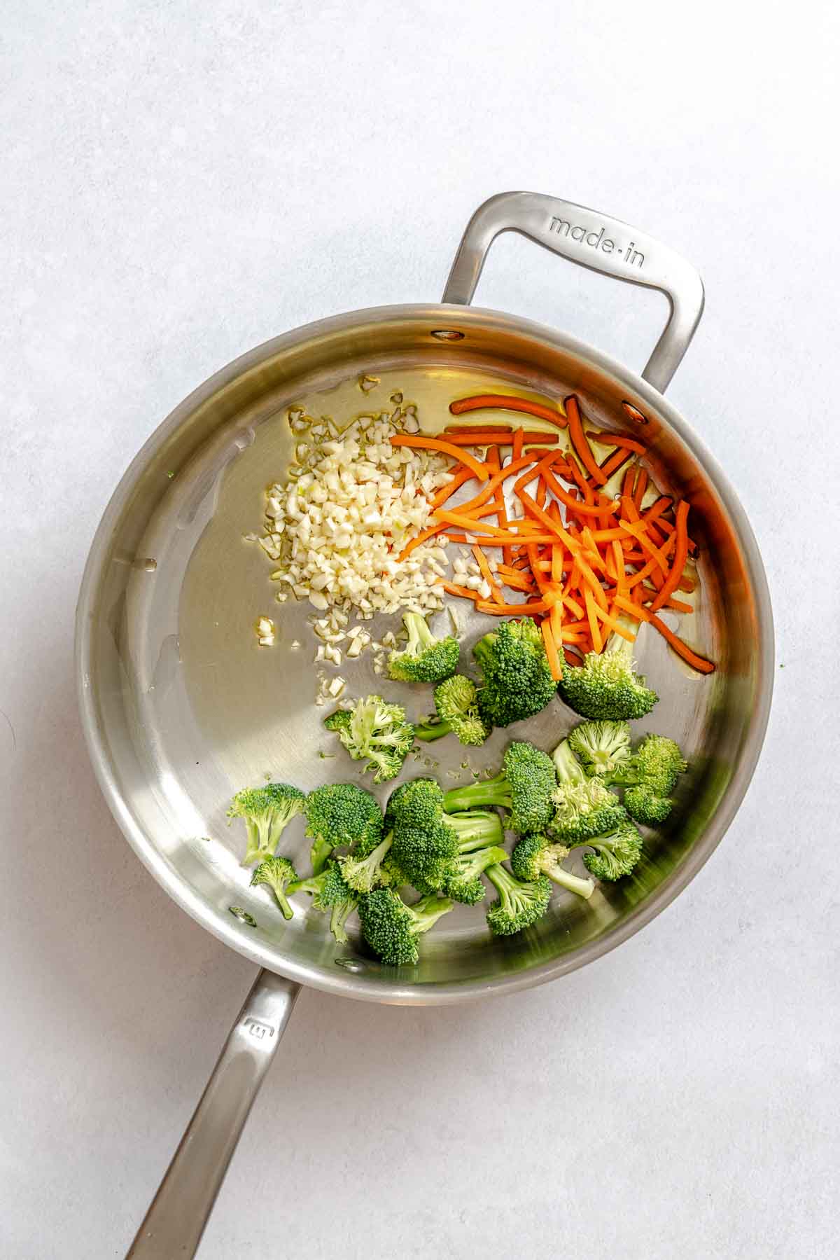 Raw broccoli, carrots, and garlic in a stainless steel skillet, ready to sauté for vegan pad Thai.