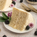 A slice of vegan vanilla cake on a plate with blackberries and flowers.