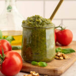 Walnut pesto piled into a glass jar sitting on a wooden cutting board with fresh basil, walnuts, and tomatoes scattered around.