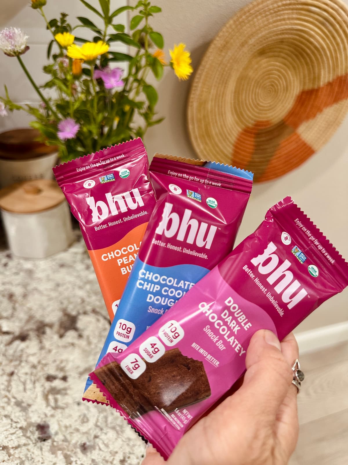 A hand holding three Bhu vegan protein bars in flavors double dark chocolate, chocolate chip cookie dough, and chocolate peanut butter.