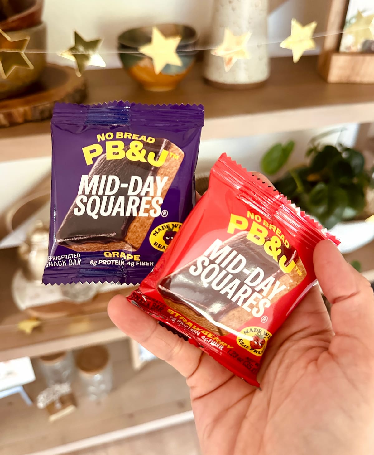 A hand holding two Mid-Day Square vegan protein bars in grape and strawberry.