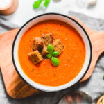 Vegan tomato soup with croutons and basil leaves in a bowl on a wooden cutting board.