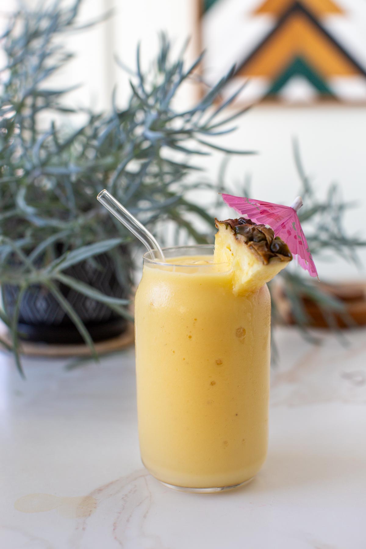 A dairy-free pineapple banana smoothie in a clear glass garnished with a pineapple chunk and a pink umbrella with a potted plant in the background. 