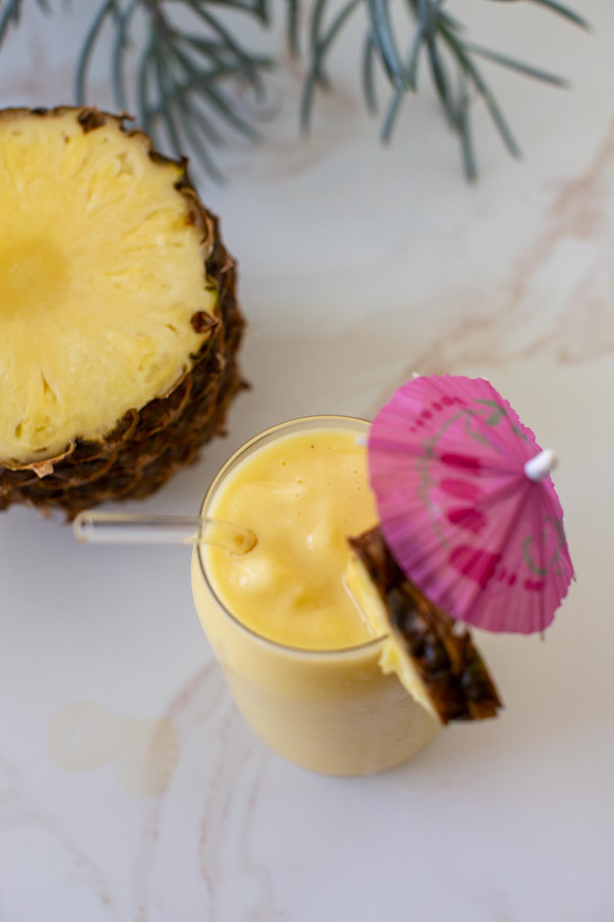 A banana pineapple smoothie without yogurt next to a half of pineapple in a clear glass with a glass straw and garnished with a pink umbrella and pineapple chunk.