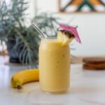 A banana pineapple vegan smoothie with pineapple slice and pink paper umbrella on white table with greenery in background.