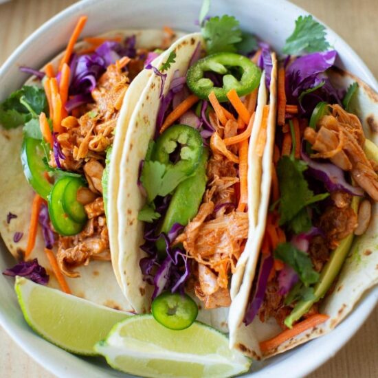 Three barbecue jackfruit tacos in a white bowl garnished with lime wedges and cilantro.