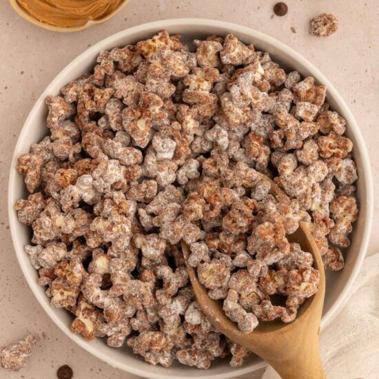 A bowl of peanut butter chocolate popcorn with a wooden scoop in it next to a small dish of peanut butter and scattered dairy-free chocolate chips.