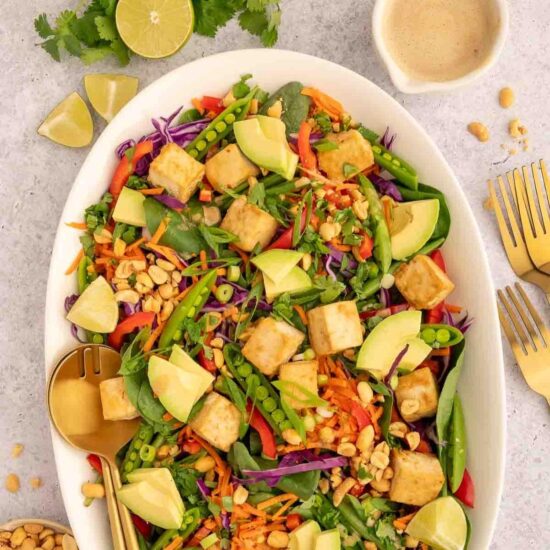 A large white bowl filled with Thai tofu salad and surrounded by lime wedges, peanut butter sauce and gold forks.