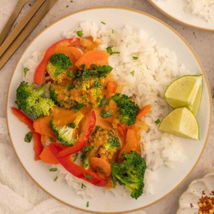 Vegan panang curry with rice, broccoli, bell peppers, and lime wedges on a white plate.