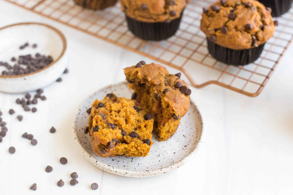 Freshly baked vegan pumpkin chocolate chip muffin open on a plate featuring a warm, golden-brown color and mini chocolate chips scattered around. 
