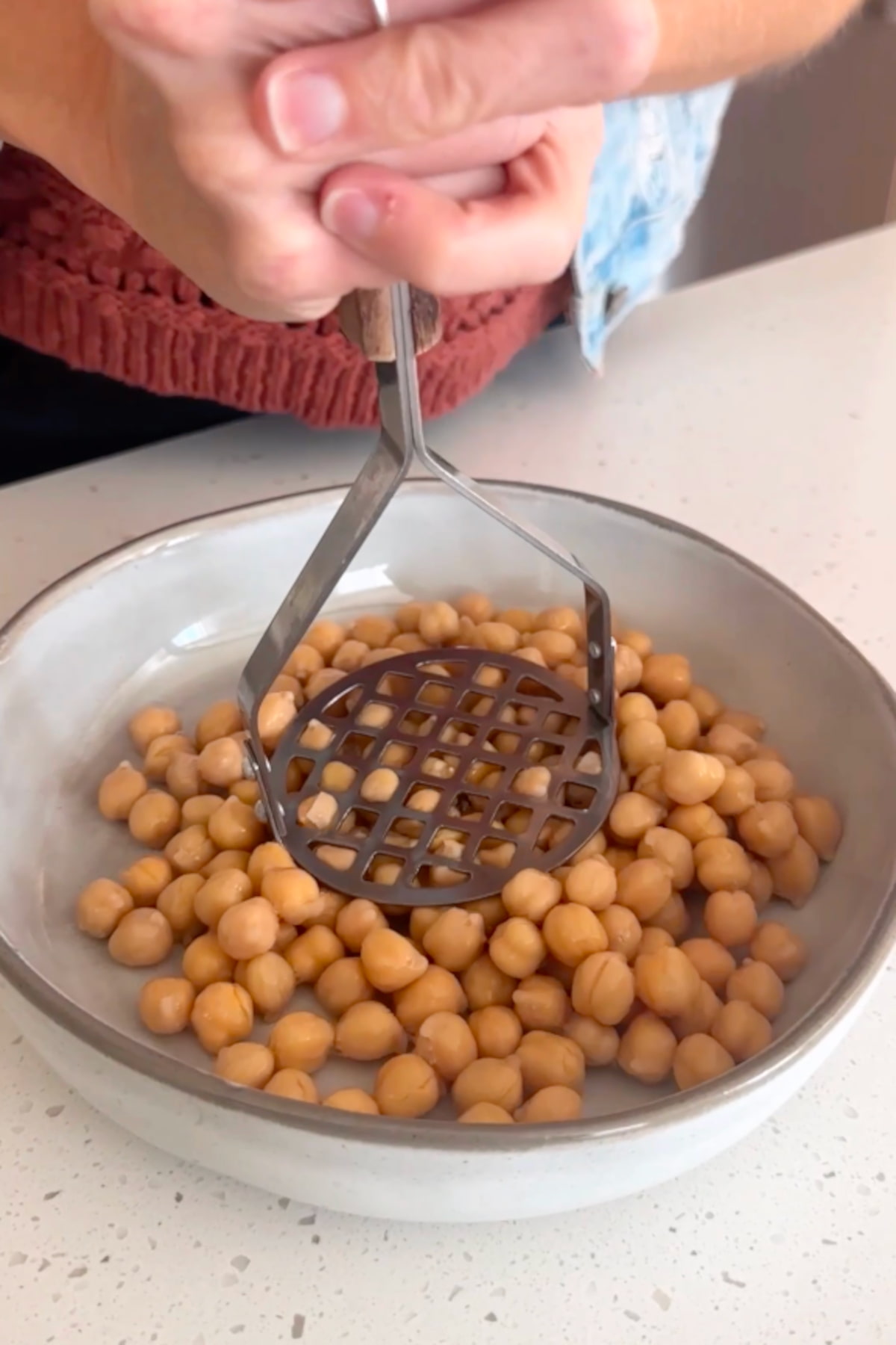 Chickpeas being pressed with a potato masher in a white bowl.