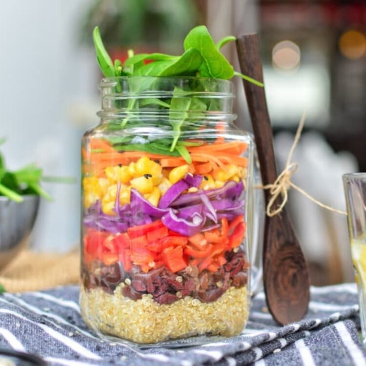 A rainbow salad recipe in a glass jar with a wooden spoon on top of a blue cloth napkin.