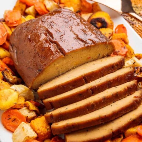 A homemade loaf of seitan turkey on a white platter surrounded by roasted vegetables.