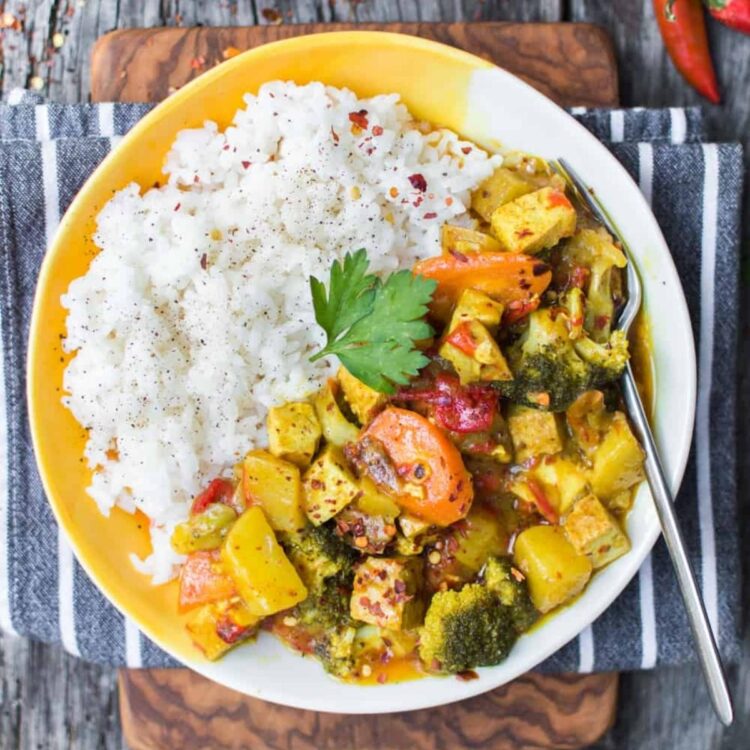 A serving of vegan yellow curry recipe along with steamed white rice served on a white and yellow plate.
