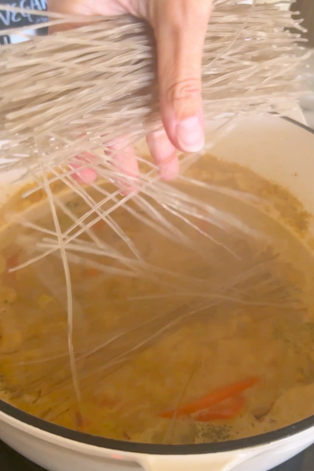A hand dropping glass noodles into the simmering soup in a large pan.