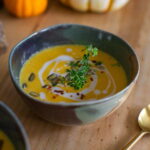 Vegan butternut squash soup garnished with cashew cream and fresh greens next to a gold spoon.