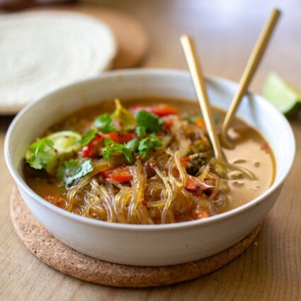A ceramic bowl full of homemade glass noodle soup on a cork trivet with gold-toned utensils.