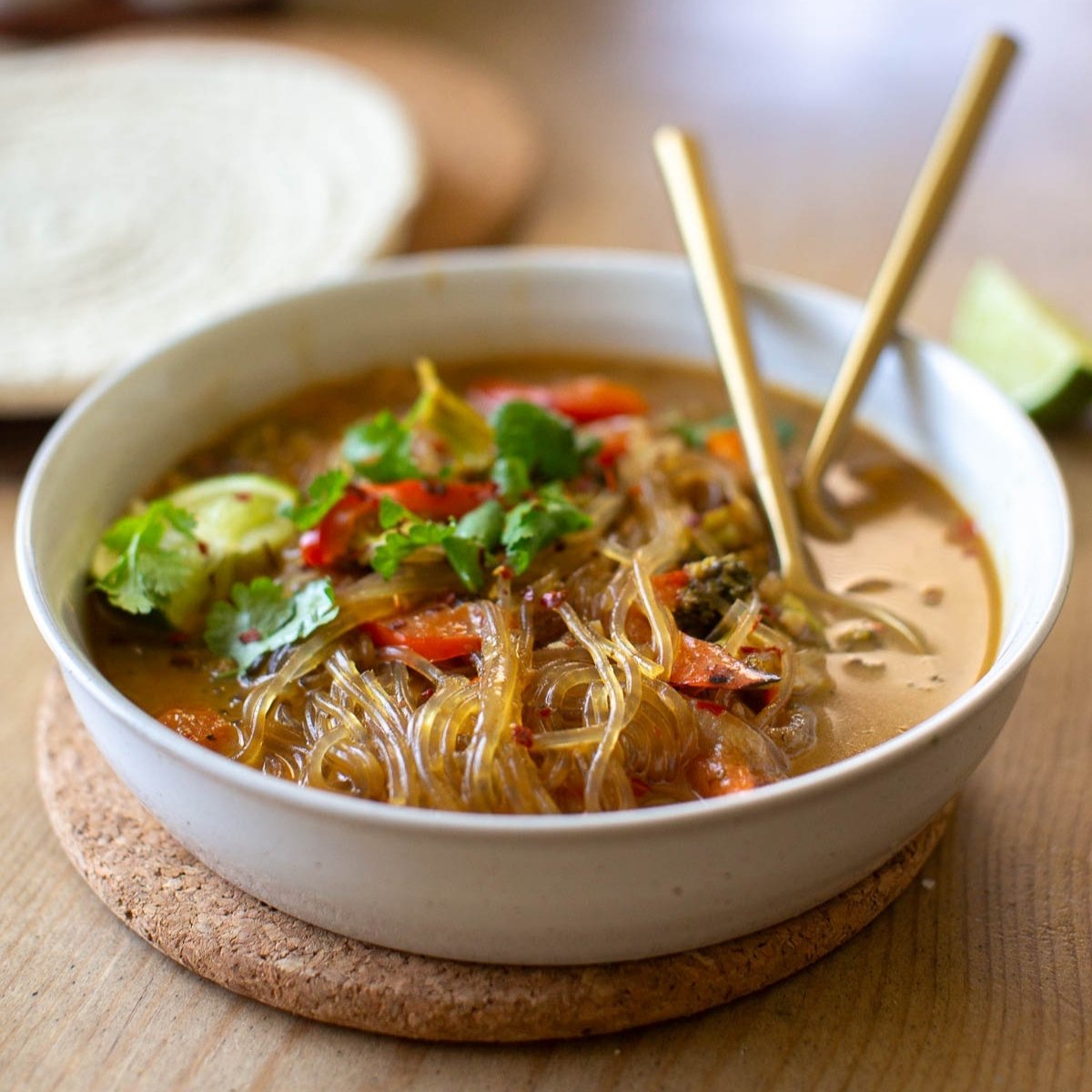 A ceramic bowl full of homemade glass noodle soup on a cork trivet with gold-toned utensils.