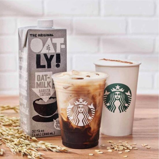 Vegan options at Starbucks including a container of Oatly plant milk, a Starbucks cold drink and a Starbucks hot beverage on a wooden countertop.