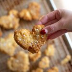 A hand with heart shaped nails holding a piece of tofu vegan fried chicken in a heart shape above a baker's rack of cooling fried tofu.