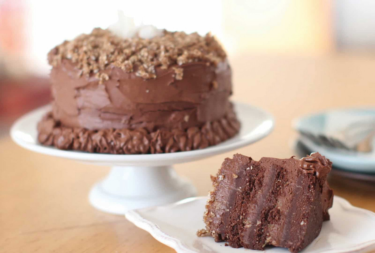 A raw vegan chocolate cake on a white serving platter next to a slice of three layer raw German chocolate cake on a white plate.