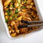 A white casserole dish with glass noodles, tofu, and gyoza dumplings layered in a savory asian sauce topped with green onions and black sesame seeds.