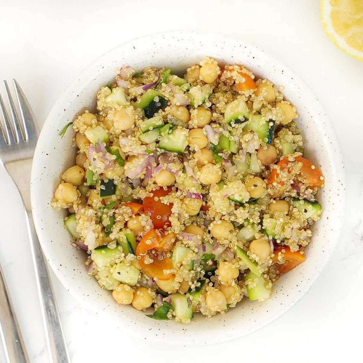 A white bowl of chickpea quinoa salad next to a fork, knife, and a half of a lemon.