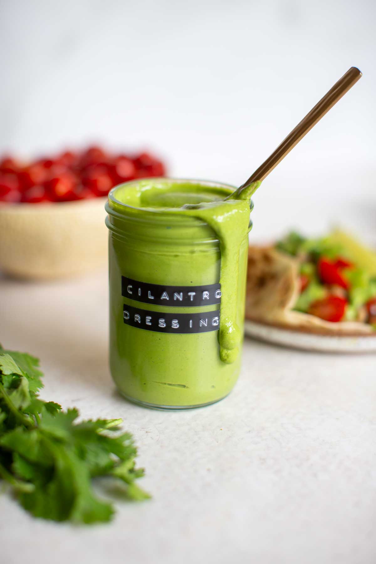 A glass jar of cilantro tahini dressing dripping from a jar with a spoon in it and a veggie taco and tomatoes behind it.