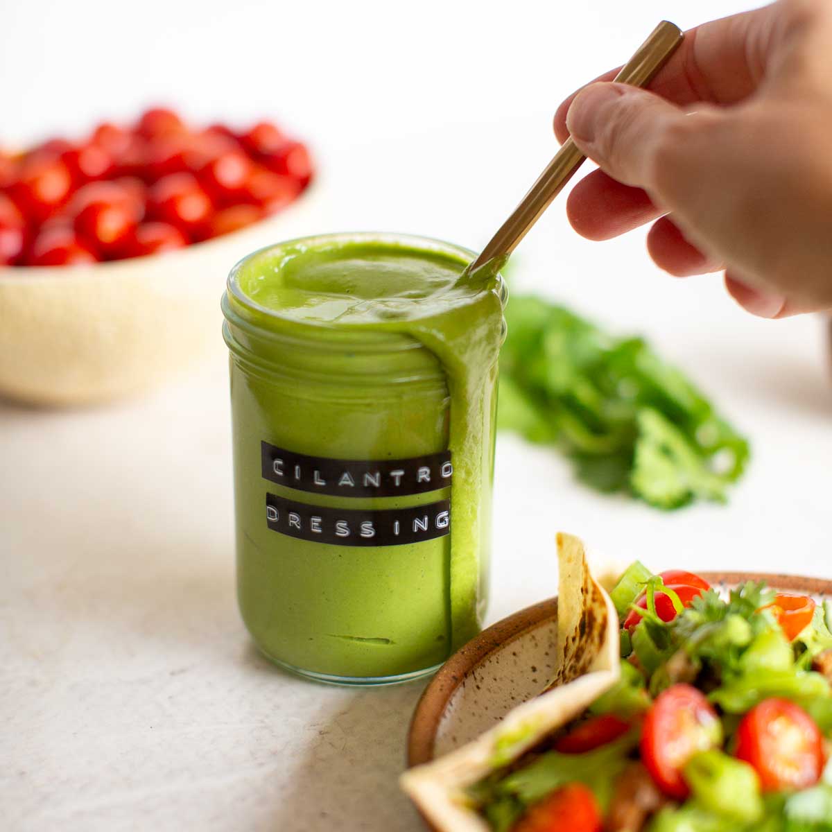 Cilantro tahini dressing dripping from a jar with a hand holding a spoon and stirring it next to a plate with a veggie taco.