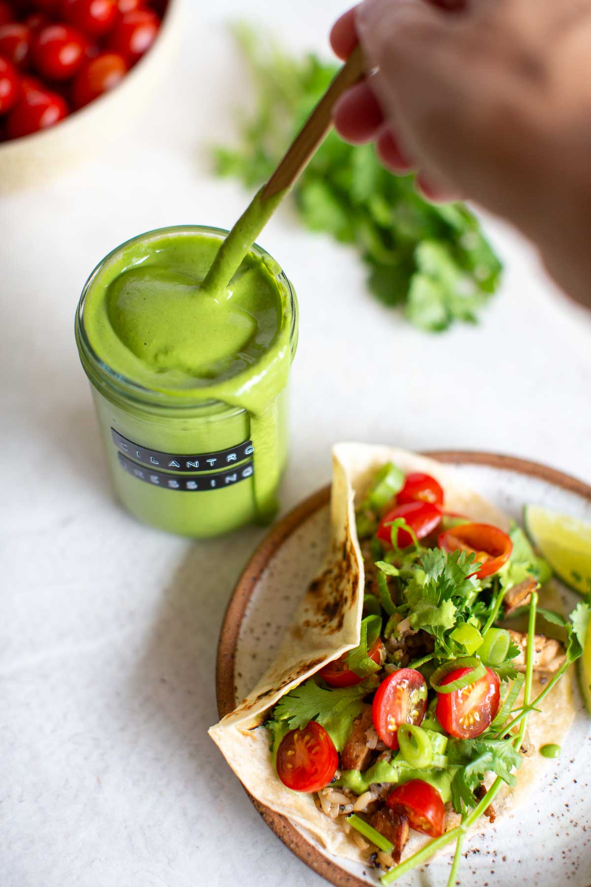 A hand stirring homemade cilantro tahini dressing next to a plate with a fresh vegetable taco with tomatoes next to it.