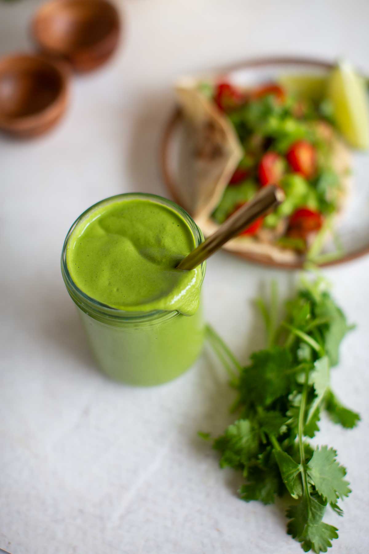 A small jar of cilantro tahini dressing with a spoon in it shot from above next to a plate with a veggie taco and fresh greens on the side.