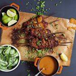 Smoky vegan seitan kebabs on a piece of parchment paper with sides of peanut sauce, lime wedges, and cucumber slices.