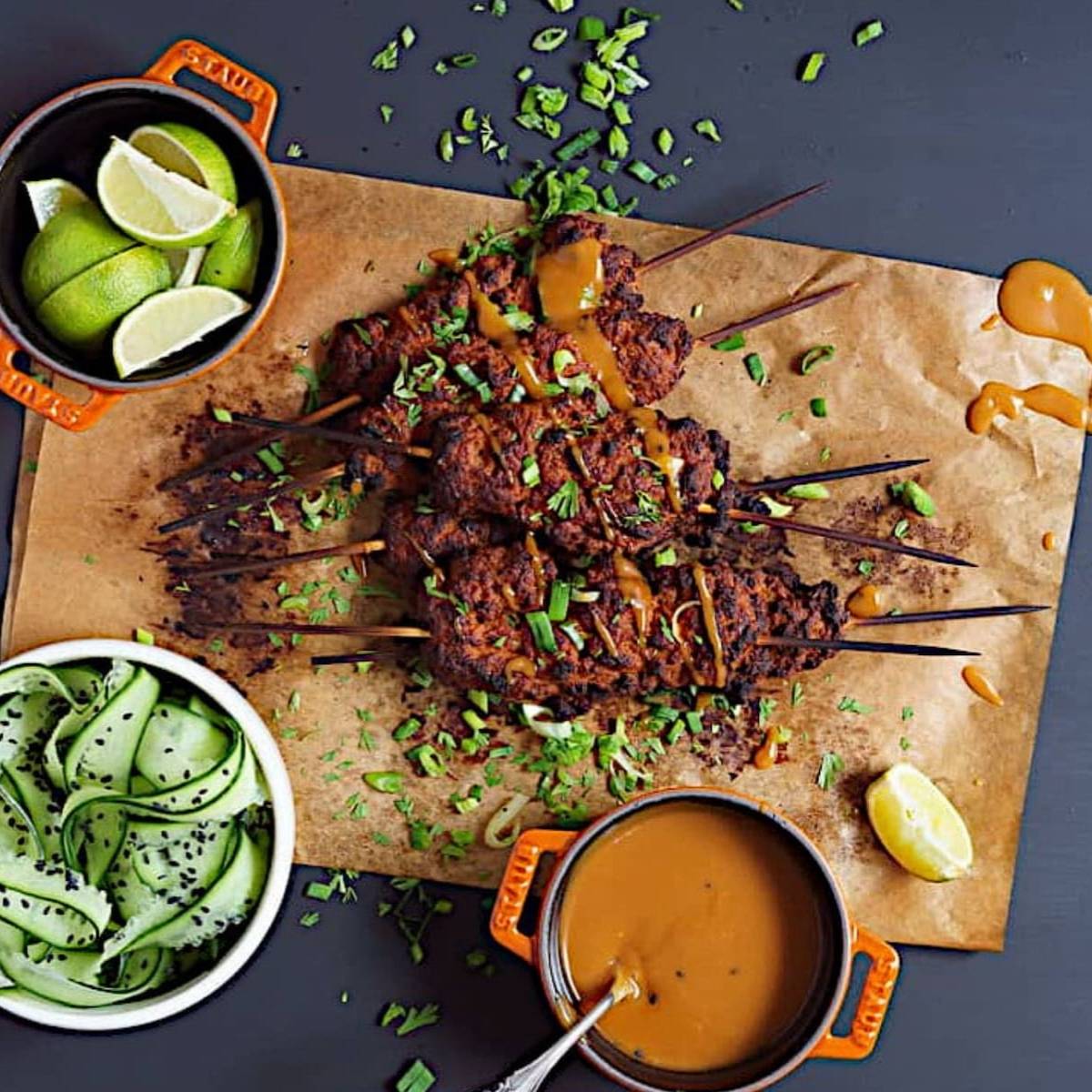 Smoky vegan seitan kebabs on a piece of parchment paper with sides of peanut sauce, lime wedges, and cucumber slices.