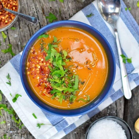 A blue bowl full of vegan sweet potato soup and garnished with fresh herbs and crushed red pepper next to a spoon and on top of a blue and white striped napkin.