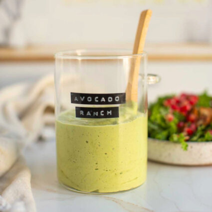 Homemade vegan avocado ranch dressing in a glass beaker with a wooden spoon next to a plate full of fresh salad.