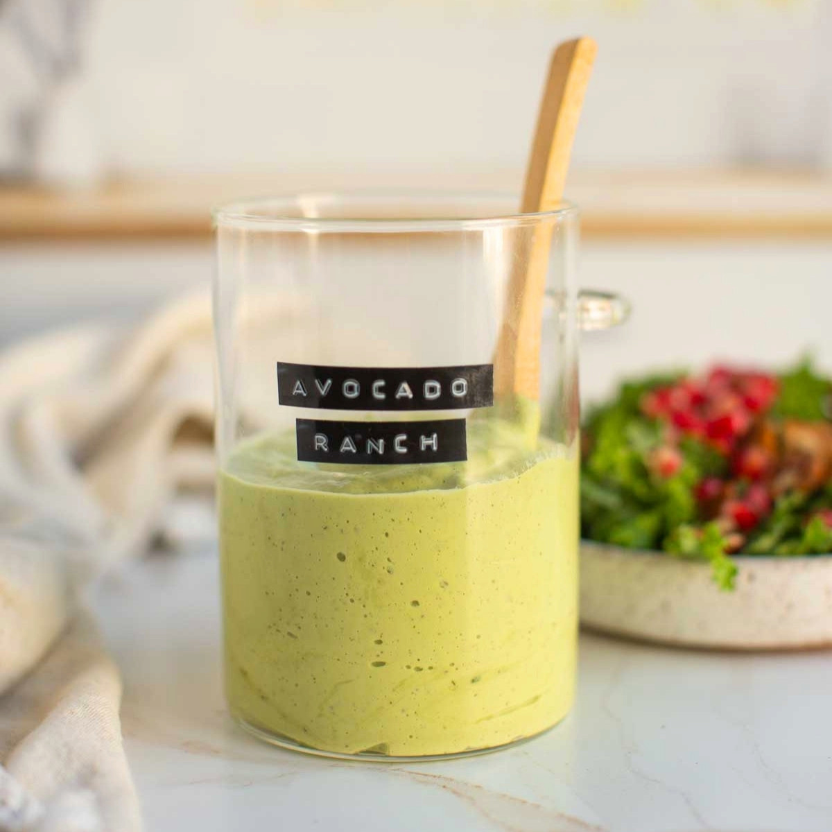 Homemade vegan avocado ranch dressing in a glass beaker with a wooden spoon next to a plate full of fresh salad.
